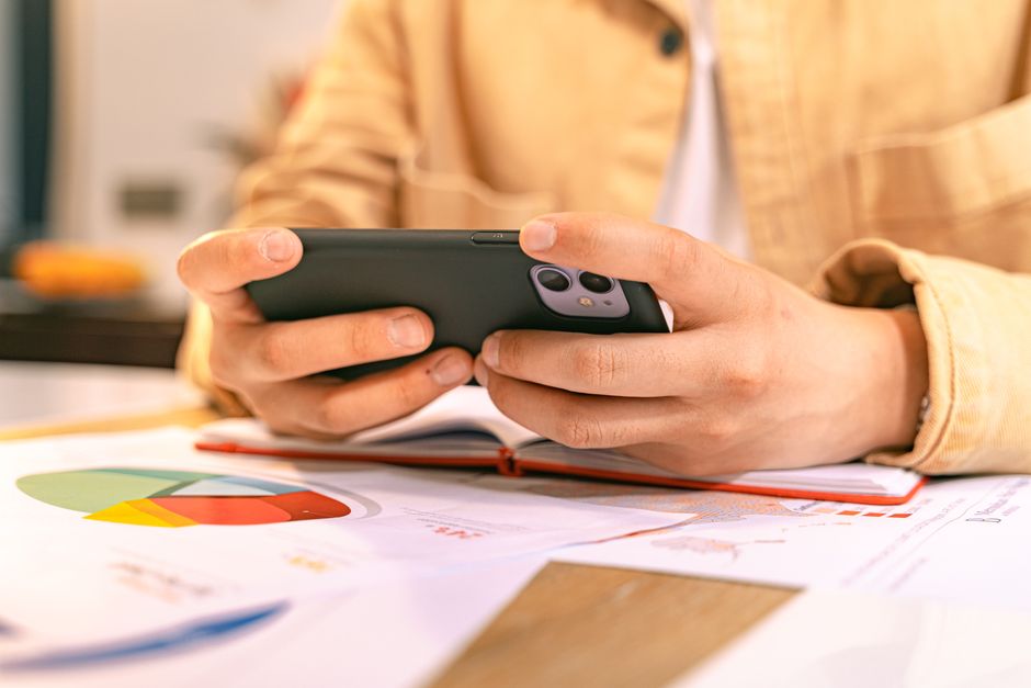 Algoritmo Redes Sociais — A business person checking their phone while working with graphs and charts on a desk.